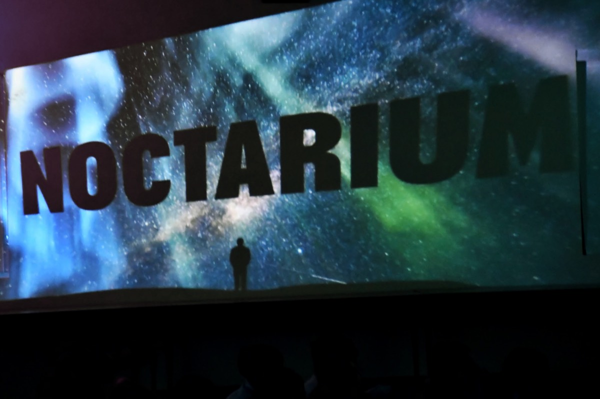The word NOCTARIUM projected in large letters across a starfield spanning a library wall.