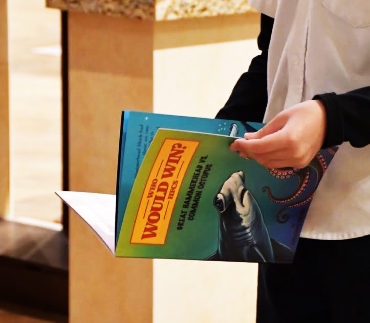 A student's hands holding an open copy of Who Would Win? Great Hammerhead vs. Common Octopus, a book from the school's own press.