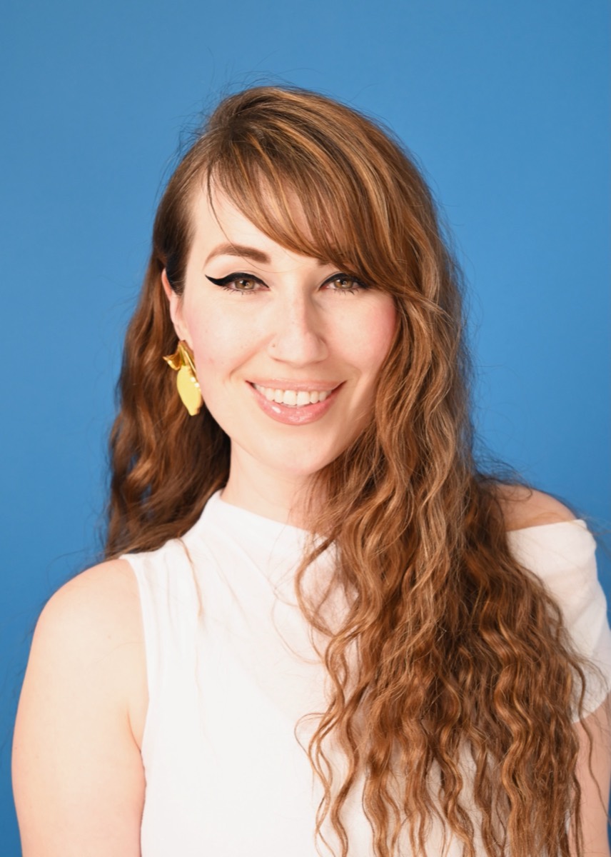 Studio portrait of Dr Verity Kahn against a blue ground, in a white sleeveless top with gold leaf earrings.